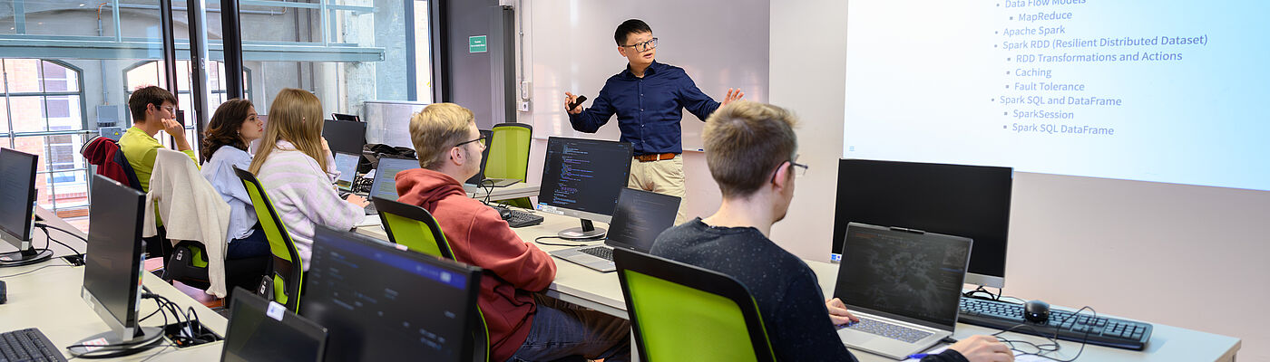 Blick in einen Seminarraum auf eine Sitzreihe mit Studierenden von hinten, vorne an der Tafel spricht und gestikuliert ein Professor. 
