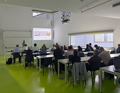 Hier ist ein Lehrraum zu sehen, in dem viele Menschen drin sitzen. Diese hören einem Vortrag zu.