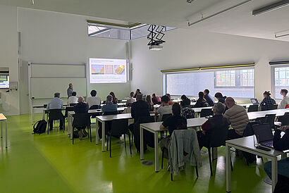 Hier ist ein Lehrraum zu sehen, in dem viele Menschen drin sitzen. Diese hören einem Vortrag zu.