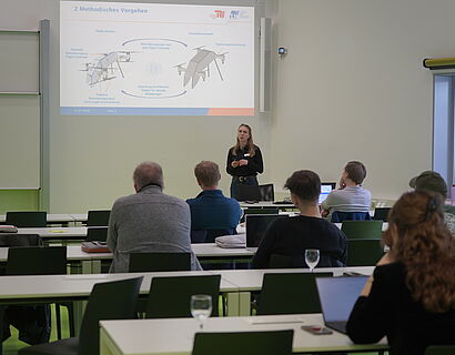 In dem Bild sieht man wie ein Vortrag von einer Person in einem Raum gehalten wird. Außerdem sitzen dort Menschen die dem Vortrag folgen.