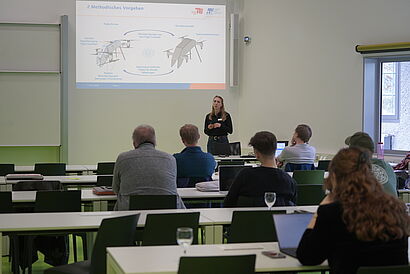 In dem Bild sieht man wie ein Vortrag von einer Person in einem Raum gehalten wird. Außerdem sitzen dort Menschen die dem Vortrag folgen.