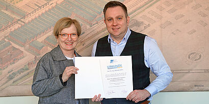 Frau und Mann stehen nebeneinander und halten eine Urkunde in der Hand.