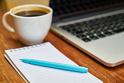 Kaffeetasse, angeschnittener Laptop, Notizblock, blauer Stift