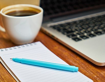 Kaffeetasse, angeschnittener Laptop, Notizblock, blauer Stift