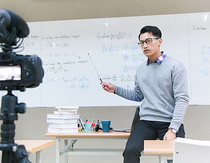 Ein junger Professor an seinem Pult vor einer Tafel zeichnet eine Lehrveranstaltung auf.