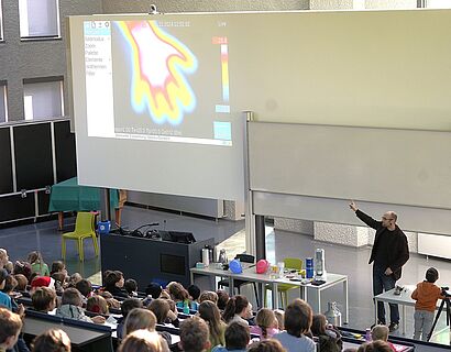 Kinderuni Winter Edition 2024 - Prof. Kleinsorge bei seiner Vorlesung. Ein Kind scannt seine Hand und diese wird auf der Leinwand mit unterschiedlichen Farben geizeigt