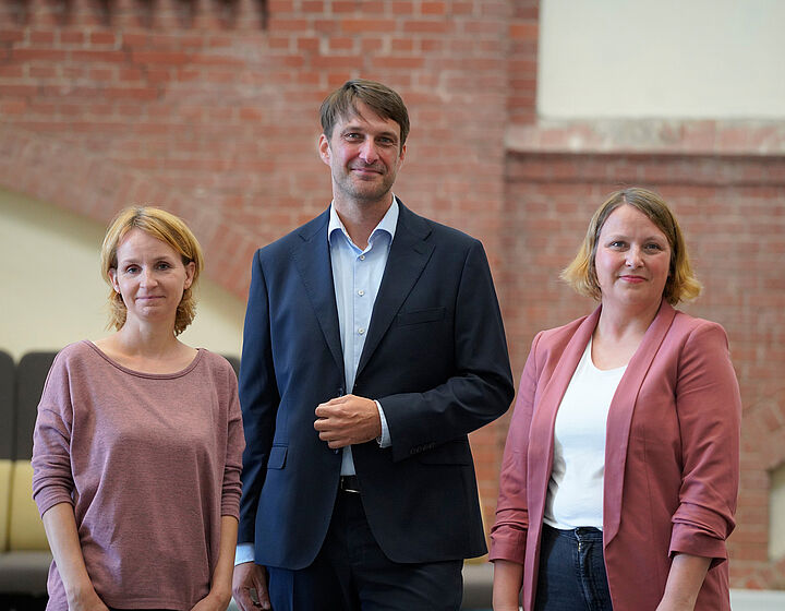 Drei Personen, Mann in der Mitte und Frauen links und rechts, stehen vor einer roten Backsteinwand
