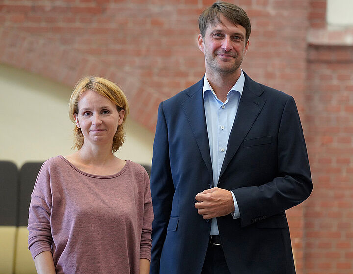 Drei Personen, Mann in der Mitte und Frauen links und rechts, stehen vor einer roten Backsteinwand