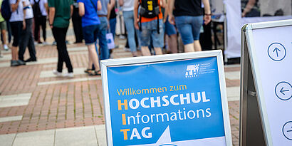 Plakat mit Aufschrift "Willkommen beim Hochschulinformationstag" auf dem Campus mit Besucher*innen im Hintergrund