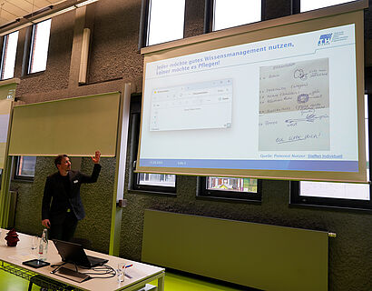 In diesem Bild ist ein Mann zu sehen, der einen Vortrag hält und dabei gerade auf seine Power Point Folie schaut.