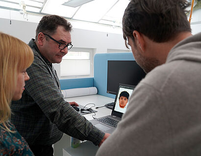 Zwei Männer und eine Frau schauen auf einen Laptop. Auf dem Laptop ist ein Phantombild abgebildet