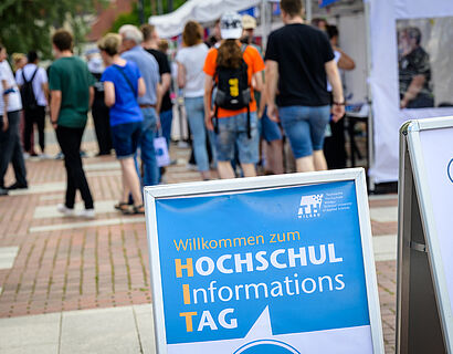 Plakataufsteller des HIT 2024 vor einer Personengruppe auf dem Campus im Hintergrund.