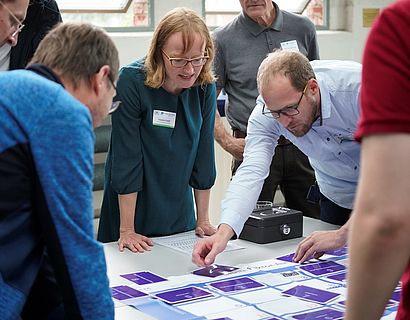 Teilnehmende des Awareness Forum 2023 stehen um einen Tisch und führen zusammen ein Serious Game zum Thema Informationssicherheit durch