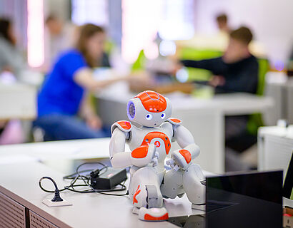 Nano-Roboter sitzt auf einem Tisch und im Hintergrund sind Personen zu sehen