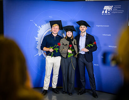 Drei Absolvent*innen stehen mit Doktohüten vor einer Plakatwand und lächeln in die Kamera