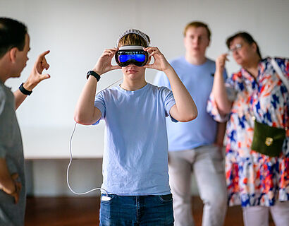 Jugendlicher probiert eine VR-Brille auf, im Hintergrund stehen Personen und schauen zu