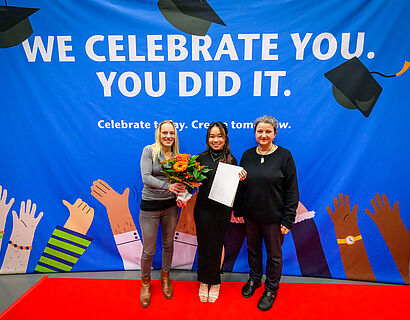 Eine Absolventin mit einem Blumenstrauß und zwei Gratulantinnen vor einer Fotowand