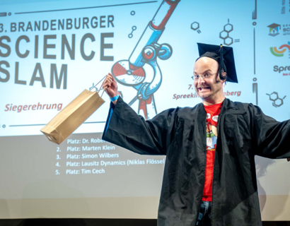Ein Mann mit Abschlusskappe und schwarzer Robe lacht auf einer Bühne.