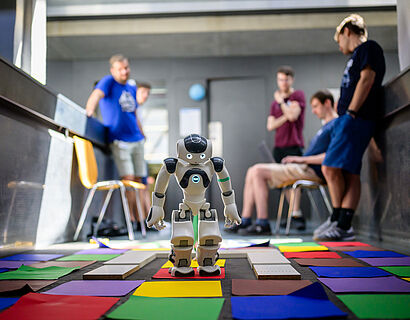 Kleiner humanoider Roboter steht auf bunten Zetteln und Platten, im Hintergrund sind Menschen und schauen zu