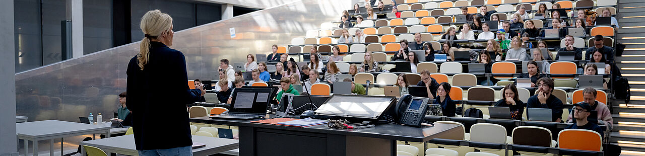 Rückenansicht einer Professorin, die in einem großen Hörsaal eine Vorlesung hält.