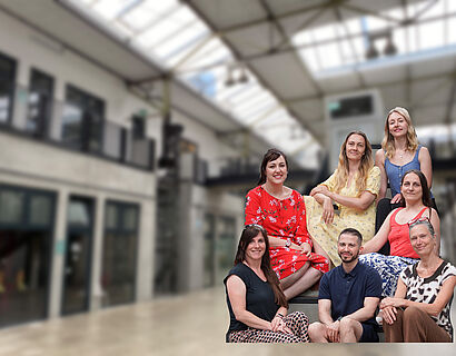 Sieben Personen sitzen für ein Gruppenbild auf einer Treppe und schauen in die Kamera
