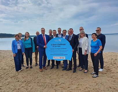 Personengruppe steht am Strand und hält ein Schild mit der Aufschrift "5-Seen-Challenge" in die Kamera