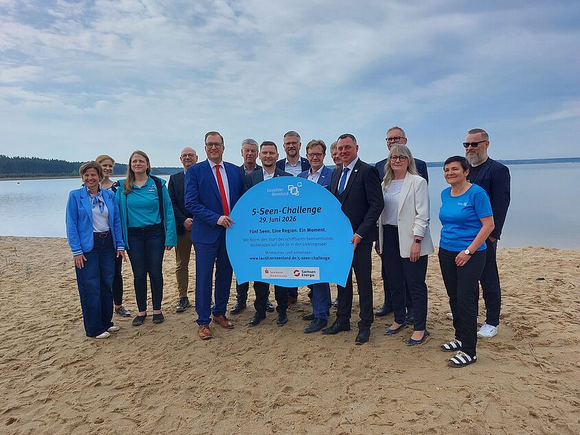 Personengruppe steht am Strand und hält ein Schild mit der Aufschrift "5-Seen-Challenge" in die Kamera