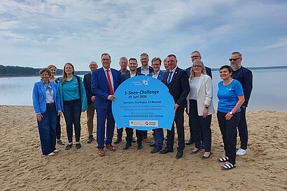 Personengruppe steht am Strand und hält ein Schild mit der Aufschrift "5-Seen-Challenge" in die Kamera