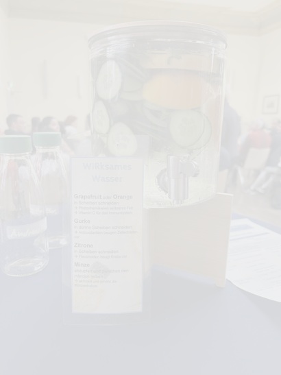 Auf dem Bild ist ein 10L Glasbehälter mit Gurke, Zitrone, Minze & Orange geschnitten im Wasser zu erkennen. Daneben ein Rezept für "WIRkrsames Wasser".