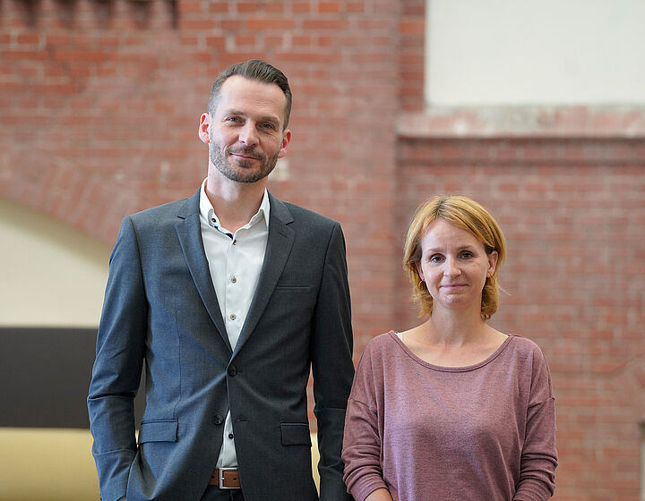 Zwei Personen, Mann links und Frau rechts, stehen vor einer roten Backsteinwand
