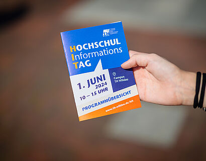 Flyer des HIT 2024 in einer Hand mit schwarzen Armbändern am Handgelenk.
