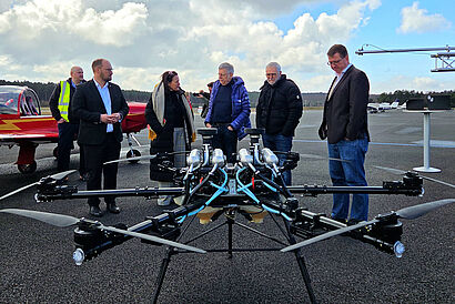 Personen stehen in einer Gruppe zusammen und unterhalten sich, im Vordergrund ist eine große Drohne zu sehen