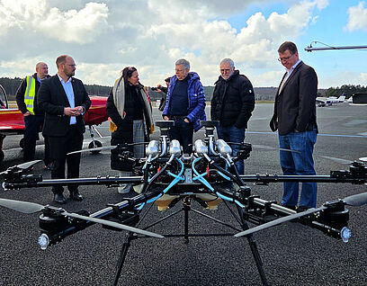 Personen stehen in einer Gruppe zusammen und unterhalten sich, im Vordergrund ist eine große Drohne zu sehen