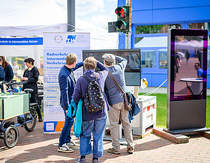 Drei Personen stehen vor einer Ampel auf dem Campus der TH Wildau und schauen auf einen dazugehörigen Bildschirm