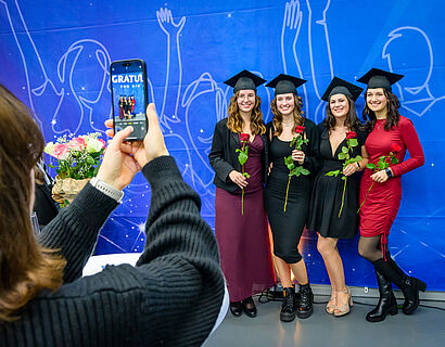 Absolventinnen stehen für ein Gruppenfoto vor blauem Hintergrund zusammen und werden von einer Person mit Handy fotografiert
