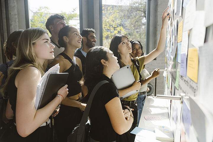 Studierende stehen in einer Gruppe vor einer Pinnwand und verschiedene Personen zeigen mit ihren Fingern auf die dort angebrachten Zettel
