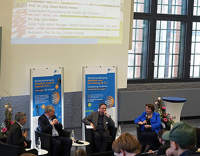 Personen sitzen in einer Gesprächsrunde mit Roll-ups im Hintergrund