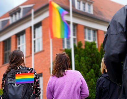 Personen stehen vor einem Gebäude und schauen auf eine gehisste Pride Flag