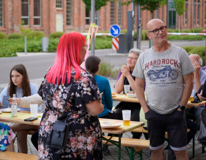 Zwei Personen stehen vor Menschen, die an Bierbänken sitzen 