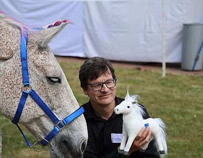 Professor sitzt neben einem Pferd und lässt es an einem weißen Stoff-Einhorn schnuppern