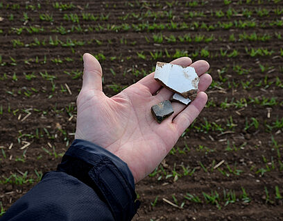 Eine Hand in Großaufnahme hält Keramikscherben in der Hand, im Hintergrund ist ein Feld zu sehen