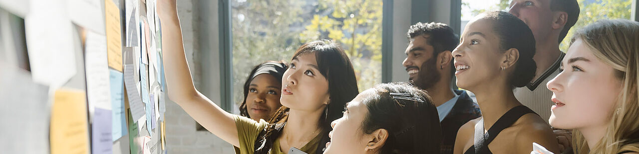Studierende der TH Wildau stehen vor einer Pinnwand und zeigen auf Inhalte
