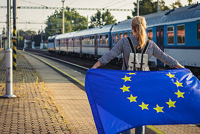 Person hält an einem Bahnhof mit vorbeifahrendem Zug eine europäische Flagge