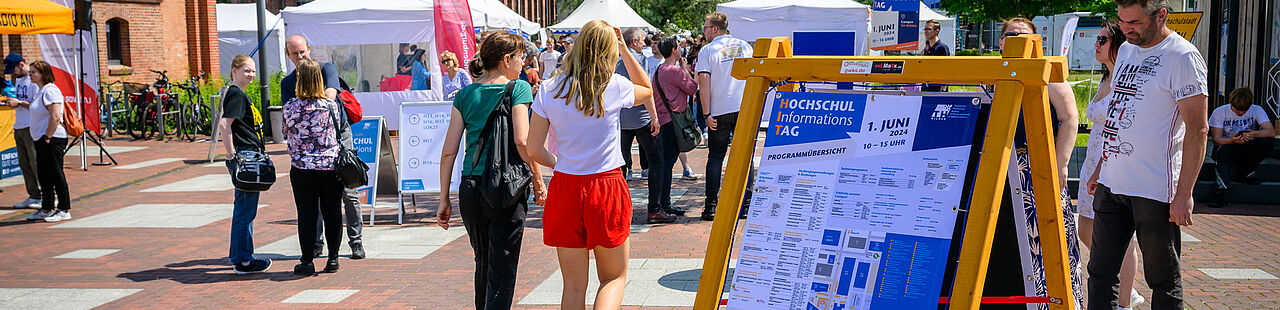 Besucherinnen und Besucher des HIT 2024 auf dem Campus der TH Wildau rund um einen großen Programmplakat-Aufsteller.