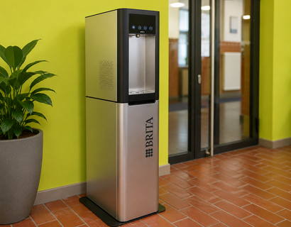 Auf dem Foto ist ein moderner Wasserspender in einem Innenbereich der Hochschule zu sehen. Er steht vor einer auffällig hellgrünen Wand, daneben befindet sich eine große Zimmerpflanze. Im Hintergrund erkennt man Glastüren, einen Flur sowie den typischen roten Fliesenboden.  Der Wasserspender wirkt sauber, gut zugänglich und zentral platziert.