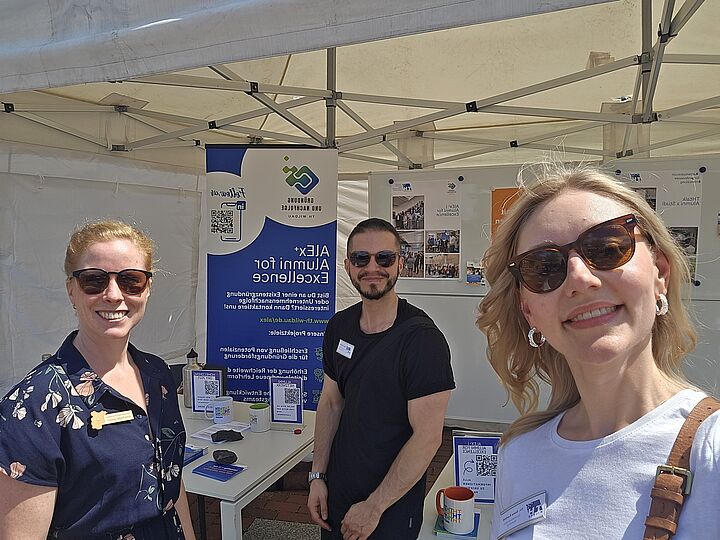 Drei Mitarbeiter*innen der TH Wildau an einem Informationsstand über die Alumniarbeit der Hochschule. Das Bild ist als Selfie von der Person im Vordergrund aufgenommen. Alle drei lächeln in die Kamera und tragen Sonnenbrillen.