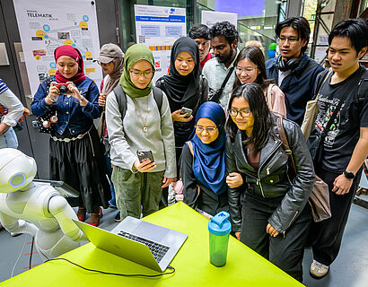 Internationale Studieninteressierte stehen um einen humanoiden Roboter herum, der auf einem Tisch sitzt