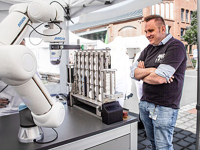 Person steht rechts neben dem Demonstrator "ViCtoR", einem Roboterarm gegen den das Spiel 4-Gewinnt gespielt wird