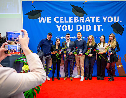 Absolvent*innen posieren für ein Gruppenbild, der Handybildschirm der fotografierenden Person ist zu sehen