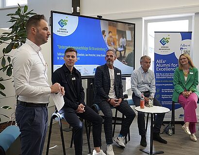 Auf dem Bild sind 5 Personen zu sehen, diese sitzen im Halbkreis vor einer elektrischen Tafel. Eine Person von ihnen steht, diese ist ganz links zu sehen und spricht in den Raum hinein.